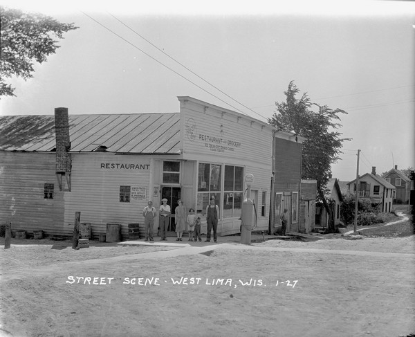 View of intersection towards a restaurant and grocery on the left, with dwellings and other buildings in the background. A group of people standing posing in front of the restaurant near a gas pump. Barrels and buckets are lined up along the side. Announcements for the circus are posted on the wall.