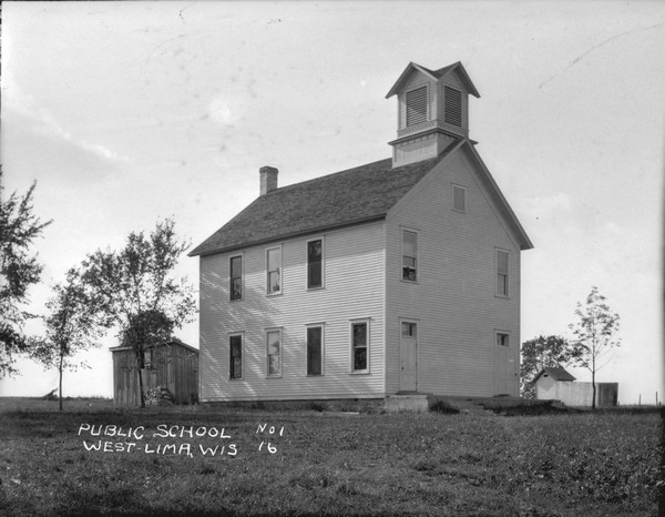 Three-quarter view from front of the public school, which is a two-story wood frame building with a belfry. There are two doors at the front of the building. A woodshed is behind on the left, and there is an outhouse on the right in the background.