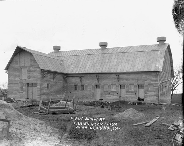 View of an L-shaped stone barn. Piles of sand, broken wood and an old plow are in the foreground.
