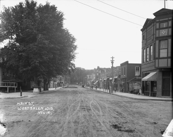 View down middle of unpaved Main Street. There are large trees along the sidewalk on the left. Businesses line both sides of the street, and there is a stable at the end of the block. On the right is a post office with a masonic lodge on the second floor. Pedestrians are on the sidewalk, and a bluff is in the background. There appears to be some street maintenance in progress on the left side of the street.