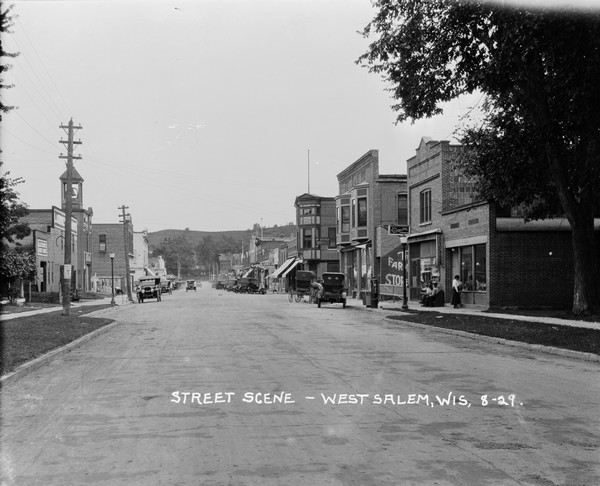 View down middle of street towards central business section. On the left is a building with a belfry. Cars and businesses line both sides of the street, pedestrians are on the sidewalk, and automobiles are parked along the curbs. There is a railroad crossing in the distance, and a bluff in the far background.