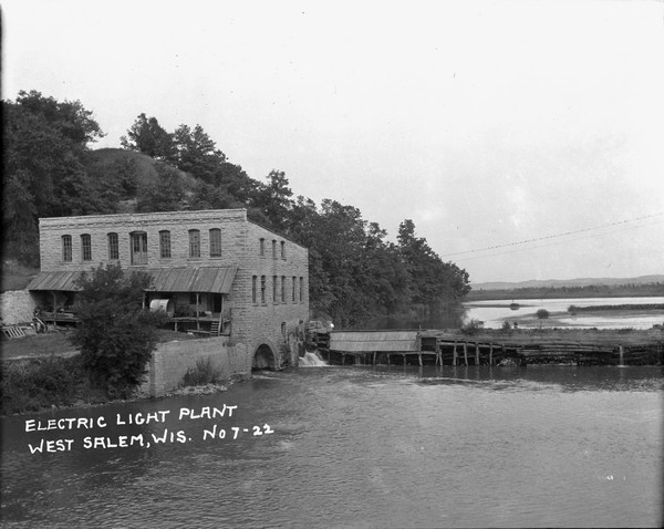 Elevated view across water of a stone building next to an earthen dam on the La Crosse River. There is an archway on the side of the building at the river level. Trees are along the banks, and there is a steep hill just behind the building. Birds are perched on power lines that run across the river. Bluffs are in the far distance.