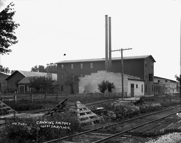 View across set of railroad tracks of a canning factory. There are a number of warehouses and outbuildings. Trees are on a hill in the background.