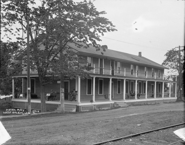 View across railroad tracks and road of the front of the Hotel May. The hotel has a wrap-around porch and balcony. Men in suits are relaxing in chairs on the porch. A birdcage is hanging on the far right end of the porch, and laundry is hanging in the back of the hotel on the left.