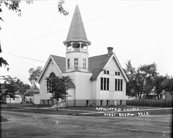 Three-quarter view from across a road of the Affiliated Church. An arched stained glass window is in the front on the left. Tall rectangular stained glass windows are along the side. A large bell is in the belfry. Young trees are planted in the lawn. There is a cornfield next door on the right.