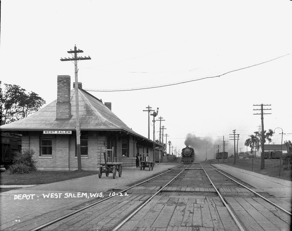 View along railroad tracks of the depot, with an approaching train coming from the opposite direction. Two men stand on the platform next to the building. Two hand carts stand alongside the tracks. Railroad cars are parked on the far left and far right on other sets of tracks.