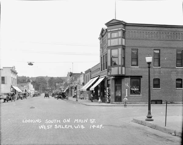 View down middle of Main Street paved with cobblestones. There is a railroad crossing at the far end, and the ridge of a hill in the far background. A barbershop and Masonic Lodge are on the corner on the right. Businesses, parked cars and pedestrians are along the street.