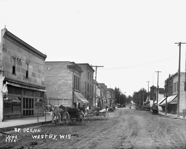 View down street in the central business district. Automobiles and horse-drawn vehicles are parked along the curbs. Businesses line the street on both sides. On the left is the Westby Farmers Alliance building, and further down the street is a barbershop pole.