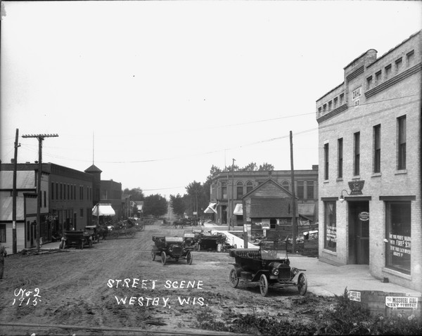 Slightly elevated view of the central business district looking down middle of street. Automobiles and horse-drawn vehicles are parked along the curbs. On the lower right is a Ford Dealership. The building has a datestone at the roofline that reads "Dahl 1913". A soda fountain and other businesses are on the left. Caption reads: "Street Scene Westby, Wis."