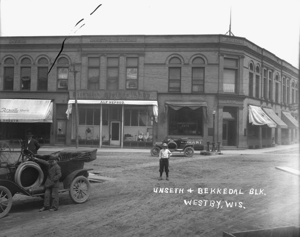 View of a street corner in a central business district. In the foreground is a boy standing next to a parked car, and another boy  and a man are standing in the street. Businesses in the background include a drugstore, clothing store and a bank. A girl is standing on the sidewalk near the bank.