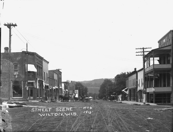View down middle of street in the central business district. A drugstore is on the corner on the left and a hotel on the right corner. Horses and wagons are parked in the street. The tree-lined ridge of a hill is in the background.