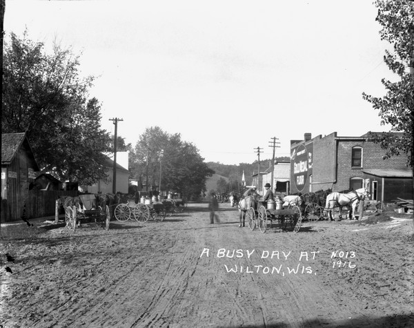 View down middle of street, with pedestrians, horses and wagons delivering milk cans. The ridge of a tree-lined hill is in the background.