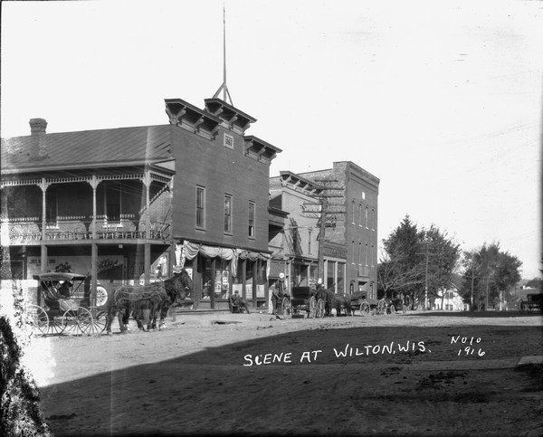 View across street in a central business district. On the opposite corner is a brick building with a balcony. A man sits in front of a store front on the side near gas pumps and an automobile. Two people are riding in a horse and buggy on the left. The horses are wearing fly-nets.