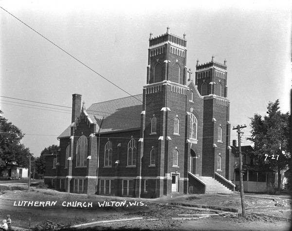 Three-quarter view of front and side of the Lutheran church. There are two belfries flanking the steps to the entrance. The main windows are arched stained glass windows. Dwelling are in the background. Two small children are in the foreground on the left. Planks of wood are laid across the muddy road.
