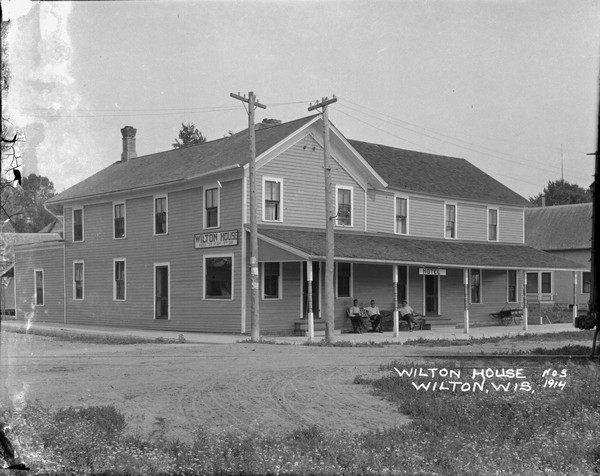 View across road and railroad tracks towards the Wilton House Hotel. A portion of the back of a railroad car is on the right. Three men are sitting on the porch of the hotel. A sign on the side of the hotel has room rates posted.