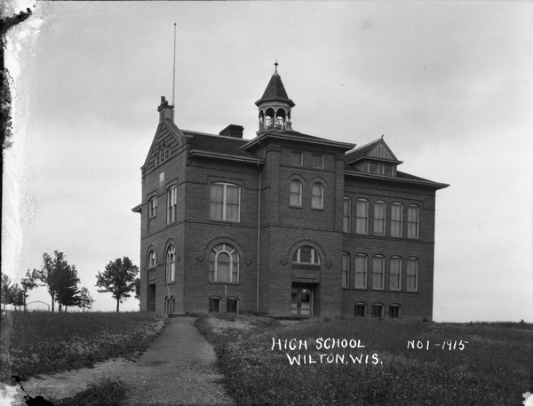 View of front and part of left side of the high school. The large brick building features arched windows, a belfry and a flagpole on the roof.