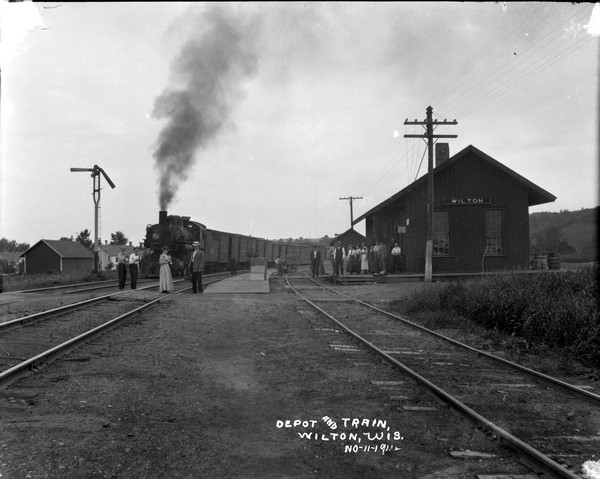 View looking down between railroad tracks towards the depot with an oncoming freight train on a third track on the far left. A group of people are standing and posing along the tracks and on the platform.