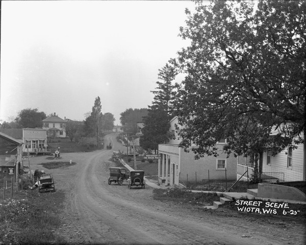 View downhill towards a central business district. A restaurant and ice cream parlor are on the right side of the road, and a general store and gas pump are further down the hill on the left. A wagon with horses is coming up the street towards the general store, and cars are parked in front of the restaurant.