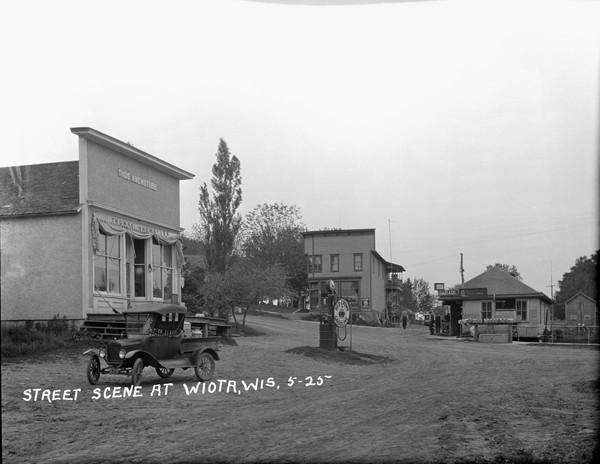 View of an intersection in the central business district. On the left is the Thomas Knewstubb  general store, with a gas pump for Red Crown Gasline out in front. A truck is parked on the left. Two men stand near the garage/service station across the road, with a row of mailboxes  lined up in front.
