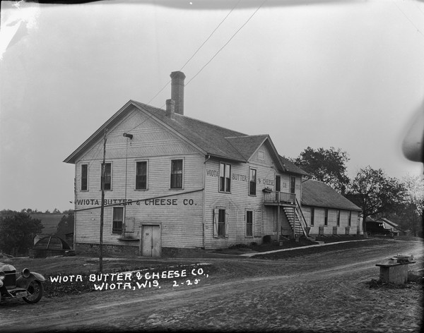 Three-quarter view of left side and front of the the Wiota Butter and Cheese Company. A wooden flight of stairs leads up to the second floor entrance. There is a double door for deliveries on the left side. Mailboxes are on a bench in the right foreground on the side of the road. A car is parked on the left.