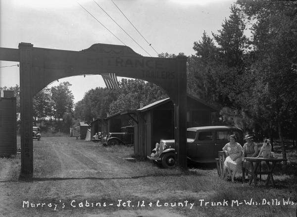 The main entrance to the Murray's Cabins Resort. On the right, two women are sitting in chairs next to a dog sitting on top of a table. The arched gate entrance has a flag hanging in the middle. Cars are parked next to small cabins among trees behind the gate.