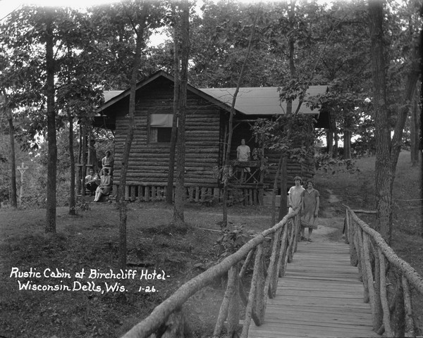 View from wooden bridge of a log cabin in the woods towards group of people. Two women standing at the end of the bridge, another woman stands on the front landing, and a man and three women pose on the back stoop. A power line is in the background on the left.