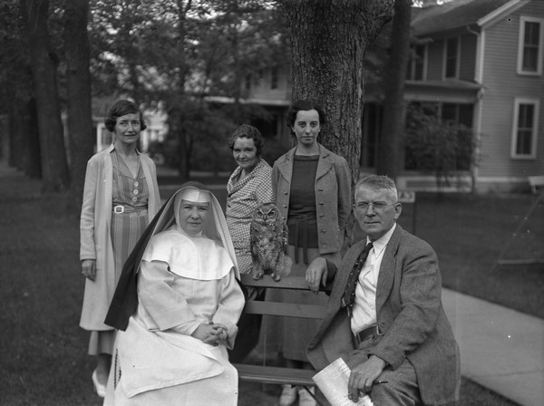 A man, a nun sit on a bench with an owl perched on the back of the bench between them. Three women stand behind them on the lawn of a resort.