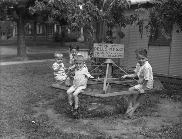 Group of four children sitting on a merry-go-round on the lawn of a resort. In the center of the merry-go-round is a sign that reads: Made by Dells Mfg. Co. See Mr. Fitzgerald." Cottages are in the background.