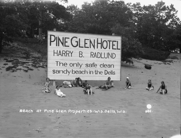 View of a beach with a group of sunbathers posing around a large billboard that reads: "The only safe clean sandy beach in the Dells."