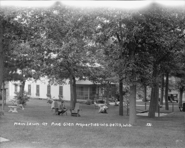 View across lawn of a group of women and one man. The guests of the hotel are relaxing under the trees on the lawn of the resort. A man and a woman are holding what may be croquet mallets. A hotel with a screened-in porch is in the background. There is a hammock and a sandbox under the trees.