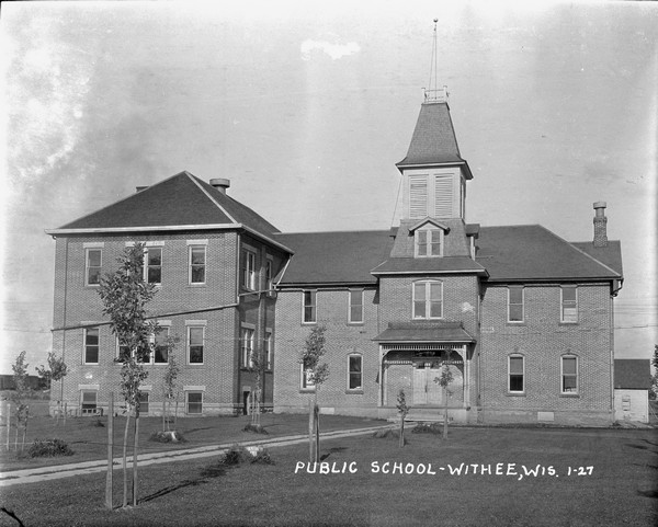 A view of the public school, a large two-story brick building with a belfry. Young trees lining the pathway to the entrance.