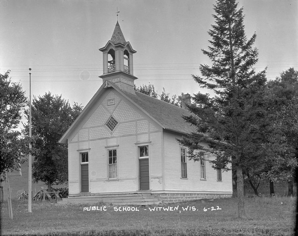 A view of the public school, a wood building with two front entrances and a belfry. A flagpole, a pile of fuel wood and a seesaw on the left. Surrounded by trees in the back.