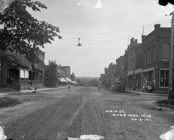 View down Center Street. The caption on the image is incorrect. On the left are storefronts, including a millinery and stable. On the right, commercial buildings include a bank, saloon and a company store. Horses and buggies are parked at the curbs. Pedestrians are walking in the street and gathered on the sidewalks. The ridge of a hill is in the far background.