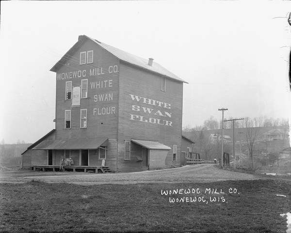 A view of the Wonewoc Mill Company where they manufacture White Swan Flour. Sacks of flour and two men sitting on the loading dock. A woman walking away on the bridge across the river. Trees and dwellings on the hillside.