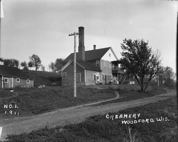 A view of the creamery and a residence on its second floor. A woman and two children at the foot of the stairs. A one lane road winding up the hill.