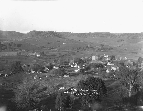 Elevated view of Woodstock, with dwellings, farm buildings and a church. Pastures and fields surround the town.