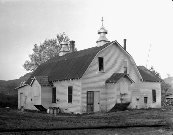 Exterior three-quarter view of white barn. There are milk cans on the left side of the building. The front and side of the barn has small extension rooms with pulleys above shuttered openings. Two large barn vents are on the roof with two weather vanes.