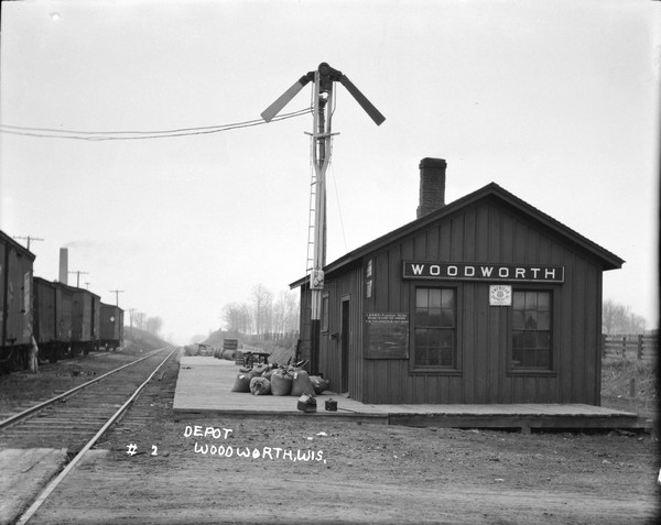View along tracks towards the depot with a tall train order signal near the doorway. A chalkboard schedule is posted near a window. Luggage and a pile of sacks are on the platform. Freight cars on another set of tracks on the left, with someone standing nearby.
