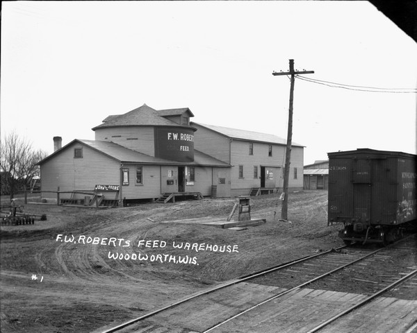 Slightly elevated view of a feed warehouse at the railroad yards. There is a truck scale with the word "Chicago" in front of the warehouse. A freight car is on one of the sets of tracks in the foreground. Signs advertise Pillsbury and John Deere.