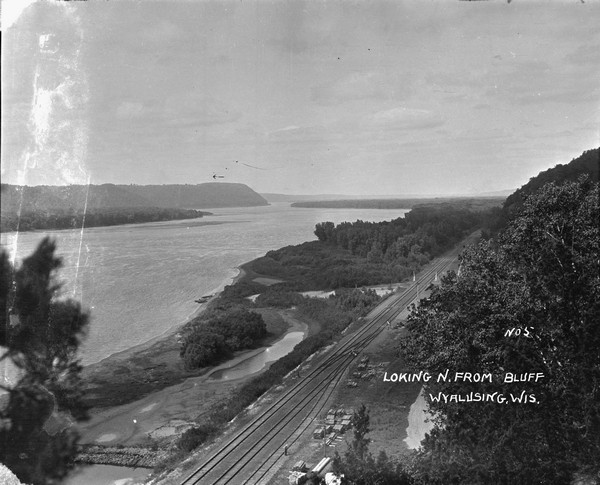 Elevated view from bluff overlooking the railroad tracks and yard along the Mississippi River. A group of men are gathered on the track near stacks of lumber. The depot is partially obscured behind a tree further up the tracks. There are wetlands between the tracks and the river.