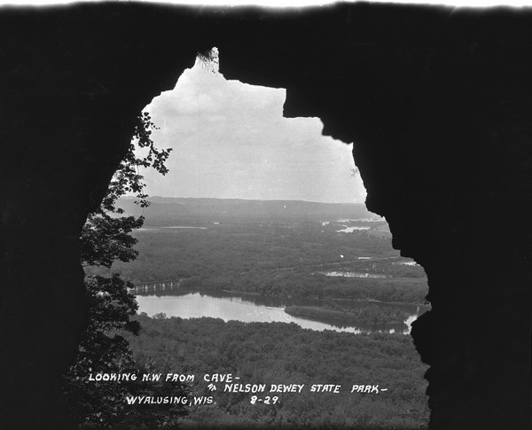 View from entrance of a cave looking out to the Mississippi River, a railroad bridge, all surrounded by a forest. There is a small island in the river.