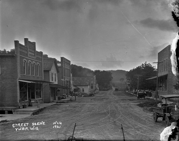 View of the central business district, with unidentified businesses, some with false fronts. Automobiles and a truck are parked along the sidewalk. A man and a boy stand at a corner on the right. There is a chicken in the middle of the street. The dirt road stretches into the distance toward bluffs in the far background.