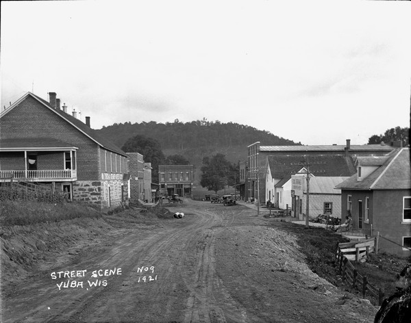 A view of the central business district. A creamery on the left. A house on the right with a woman in a rocking chair and two children in front. Some men sitting in front of a store in the background.