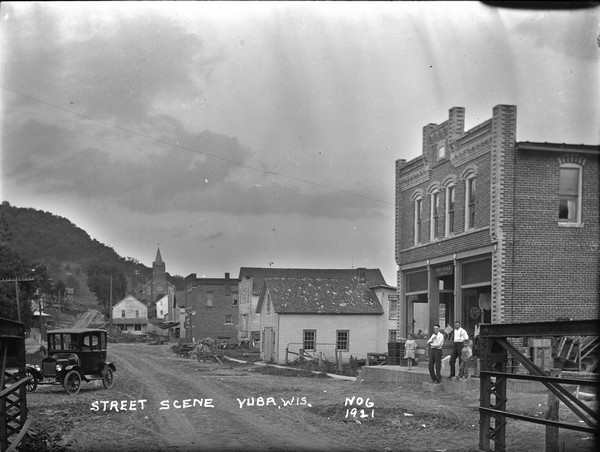 A view of the central business district from the end of a bridge. The post office on the right with a small group of men and children in front. An automobile parked on the left. A couple wagons on the right. A church in the background.