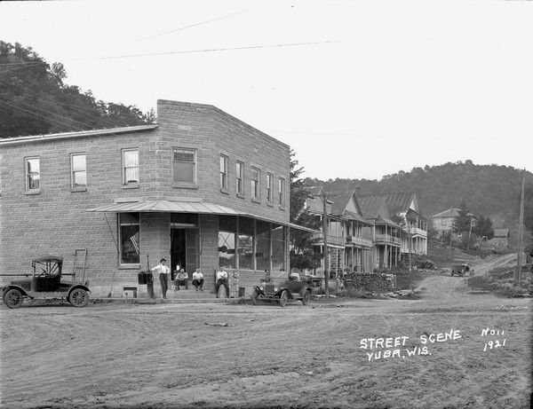 A street scene with a group of people in front of the corner store. A flag in the window. A row of houses with balconies behind that. A large stack of wood in front of the first house.