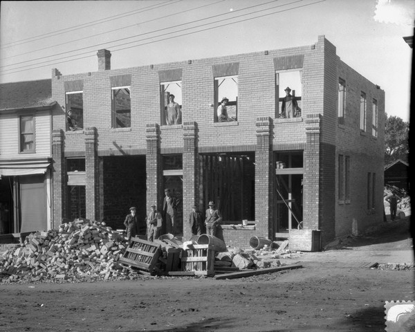 View from road of a brick building under construction. Five men are posed in front near a pile of bricks. Four men are posed in the window openings on the second floor.