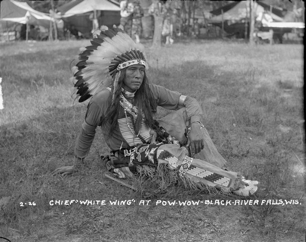 Portrait of Native American man sitting in the grass wearing headdress, beaded pants and moccasins. An axe is lying in the grass at his side.