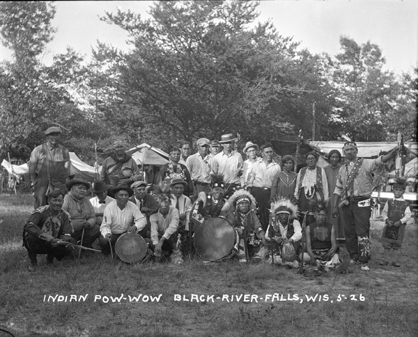 Native Americans posing for a group portrait, with two drummers are sitting in the front row.. The group is wearing a mix of western dress and native dress.