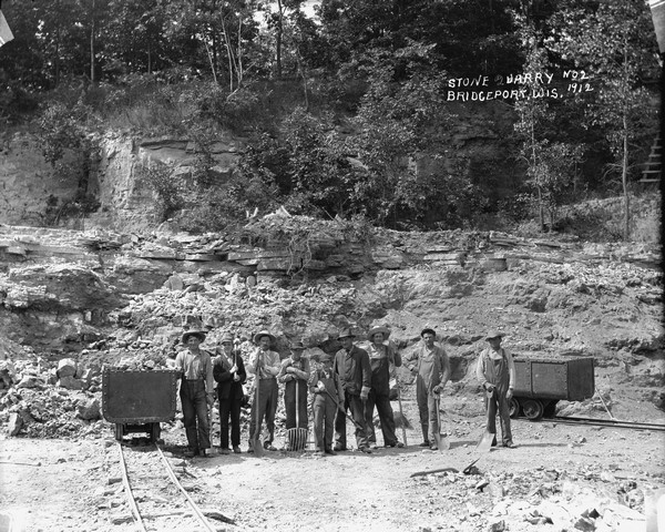 Group portrait of quarrymen posing with their implements. There are carts on tracks that lead to the cliffs of the quarry behind the group.