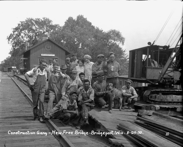 Group portrait of construction workers, two women and a dog at the railroad tracks near the depot. They are posed near a crane. A group of people stand on the platform near the depot in the background.